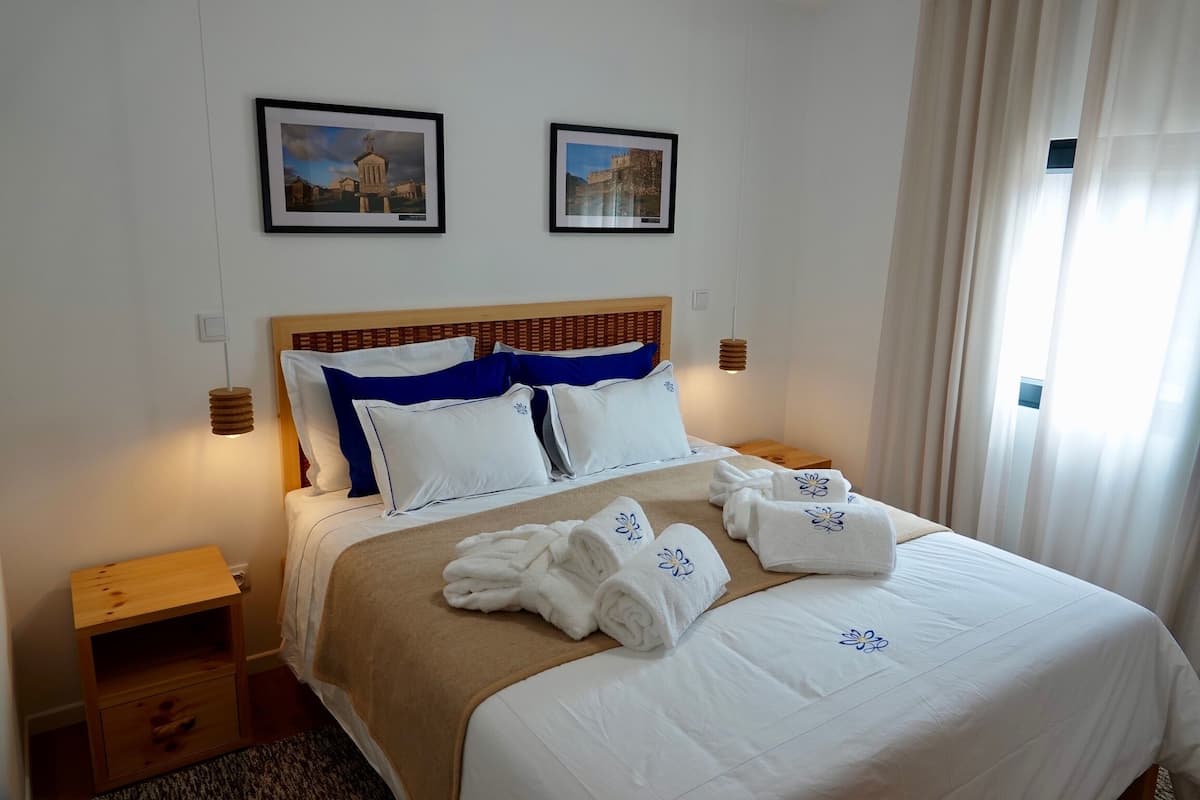 A neatly made bed with white and blue bedding, two framed pictures on the wall, and a wooden nightstand with a lamp.
