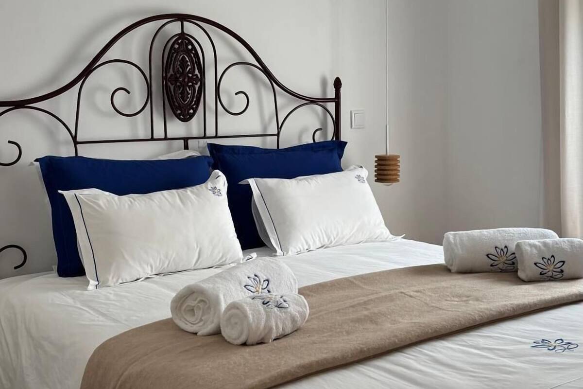 A neatly made bed with white and blue bedding, a beige throw blanket, and rolled towels.
