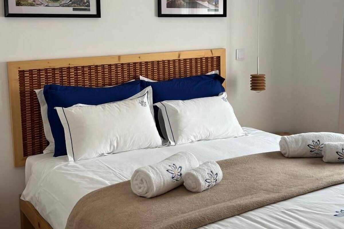 A neatly made bed with white and blue bedding, two framed pictures on the wall, and a ceiling fan.