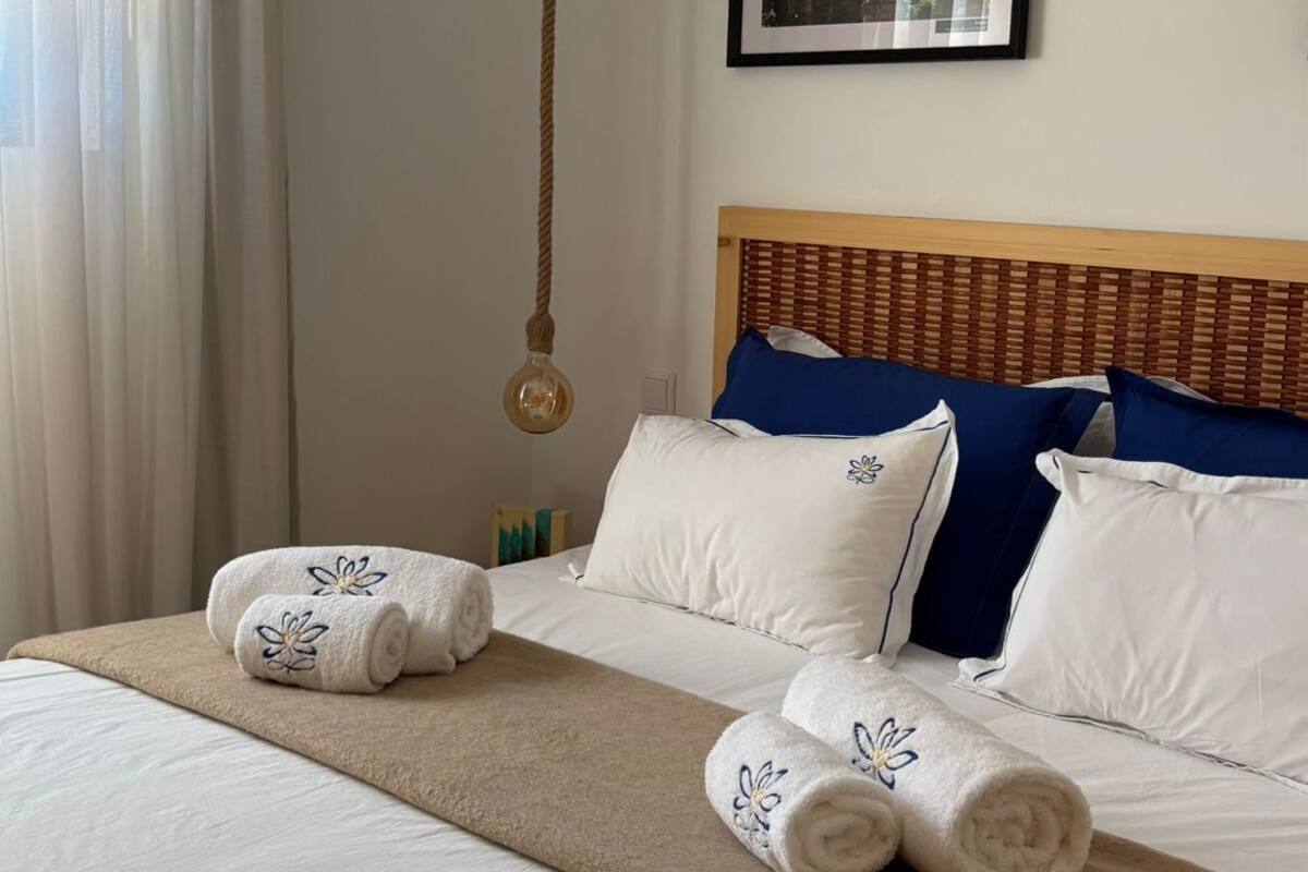 A neatly made bed with white and blue pillows, a beige blanket, and rolled towels with a floral pattern.