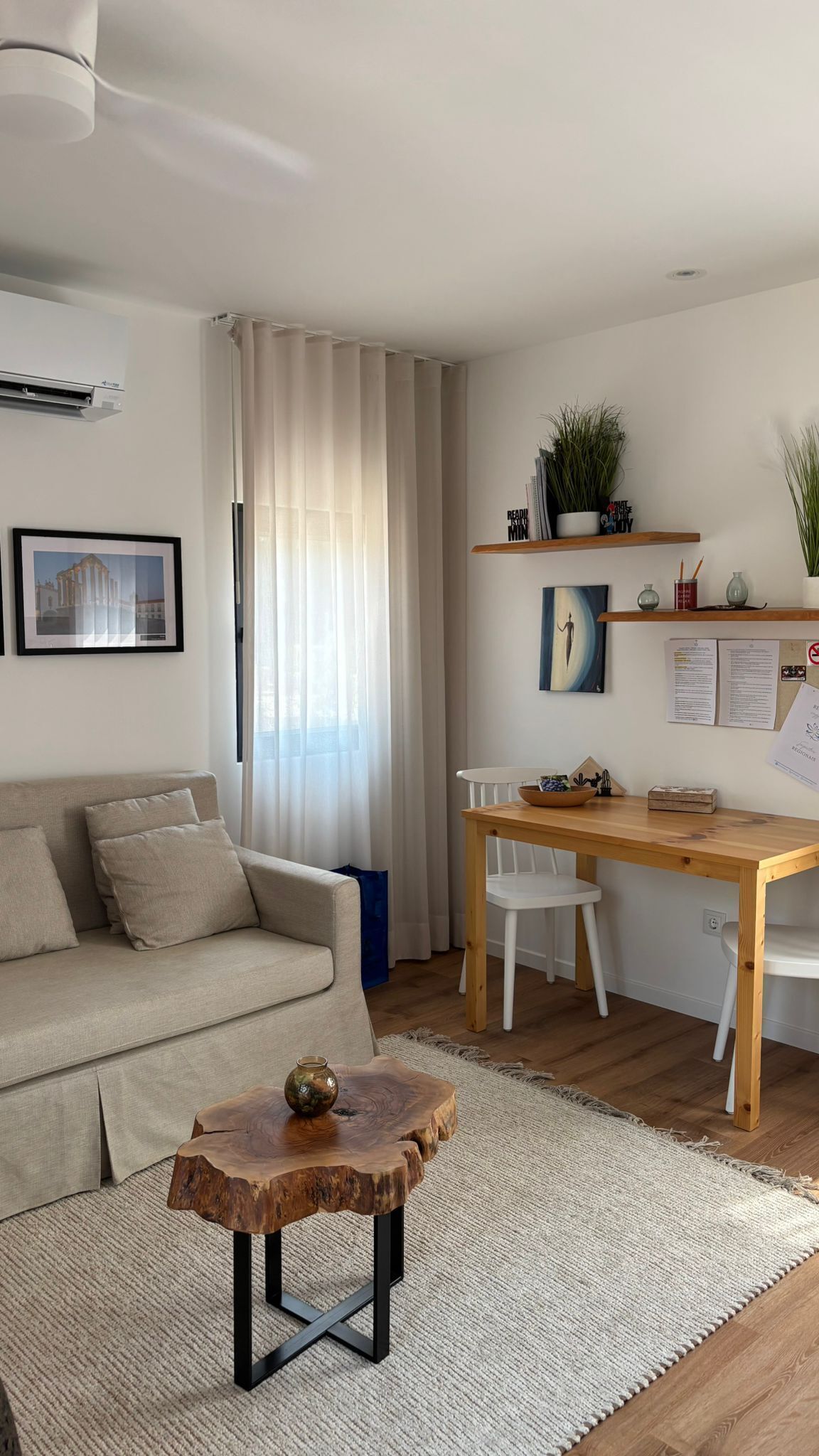 A modern living room with a sofa, coffee table, and a wooden desk with chairs. There is a window with curtains, a wall-mounted shelf with plants, and a framed picture.
