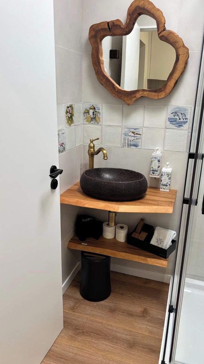 Un baño con un lavabo de piedra redonda, repisas de madera, un cesto de basura negro y un espejo con marco de madera.