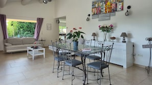 A spacious living room with a sofa, dining table, and chairs, a kitchen in the background, and a large window with curtains.