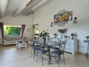 Amplia sala de estar con sofá, mesa de comedor y sillas, una cocina al fondo y un ventanal con cortinas.