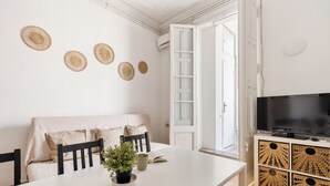 Ein modernes Wohnzimmer mit einem weißen Sofa, einem Esstisch mit Topfpflanze, einem Flachbildfernseher auf einem weißen Schrank und einem großen Fenster mit Blick ins Freie.