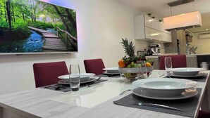 Un moderno comedor con un televisor de pantalla plana grande montado en la pared que muestra una escena de naturaleza. La habitación cuenta con una larga mesa de comedor con sillas tapizadas en rojo, un frutero y platos y cubiertos colocados con cuidado.