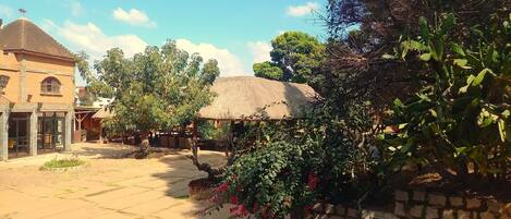 A courtyard with a thatched-roof structure, surrounded by greenery and a building with a tiled roof.