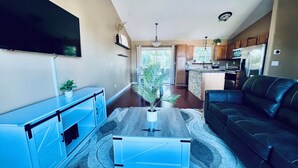 A modern living room with a flat-screen TV, a black leather sofa, a blue coffee table, and a kitchen area in the background. - NEW Spacious Home Minutes From Oneida Lake & Fall Festivities!  (Brewerton)