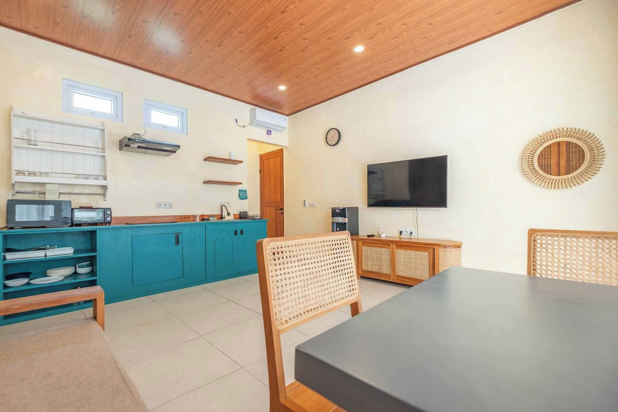 Una cocina compacta con armarios verde azulado, techo de madera, televisor negro y silla de fibras naturales.
