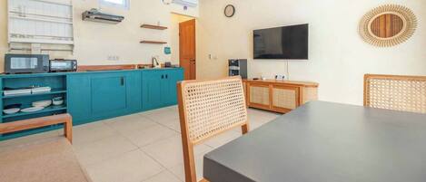 Una cocina compacta con armarios verde azulado, techo de madera, televisor negro y silla de fibras naturales.