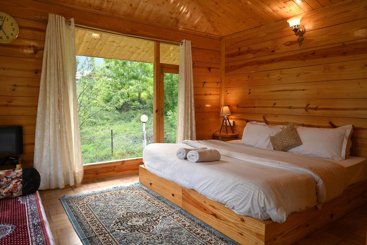 Dormitorio de cabaña de madera con cama, mesita de noche, lámpara, reloj y vistas al exterior.
