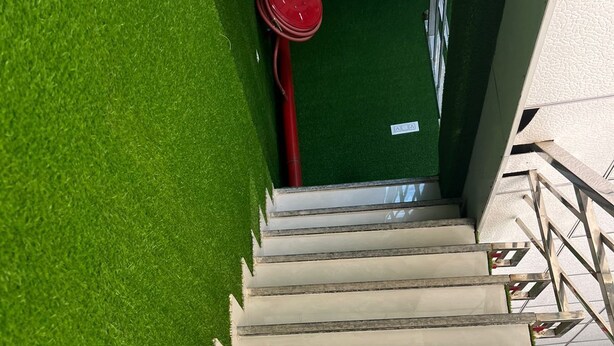 A staircase with artificial grass on one side and a white wall on the other, leading to a room with a green wall and a red object in the background. - Hotel Neki & Restaurant (Meerut)