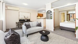 A modern living room with a grey sofa, a black coffee table, and a dining area in the background. - Executive Elegance: Stylish 1BR Suite on Hay St (East Perth)