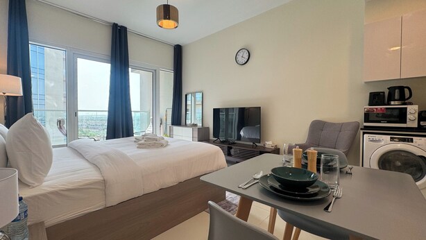 A modern hotel room with a bed, a desk, a dining table, a TV, a washing machine, and a view of the city. - Modern Studio in Virdis Damac Hills 2 (Dubai)