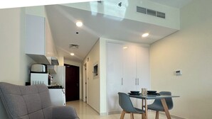 A modern kitchen with a coffee maker, a dining area with a table and chairs, and a grey armchair. - Modern Studio in Virdis Damac Hills 2 (Dubai)