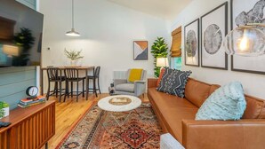 A living room with a brown leather sofa, a round coffee table, a dining area with chairs, and a TV on the left. - Steps Away Cottage • Hot Tub, Fire Pit, Downtown (Waynesville)