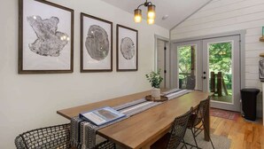 A dining area with a wooden table, black wire chairs, and framed artwork on the wall. - Birdsong Cottage | Gorgeous Lake Junaluska Retreat (Waynesville)