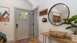 A hallway with a wooden floor, a white door, a wall-mounted mirror, a basket on a shelf, and a plant. - Birdsong Cottage | Gorgeous Lake Junaluska Retreat (Waynesville)