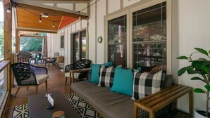 A covered porch with a wooden ceiling, a sofa with checkered cushions, and a dining area with chairs and tables. - Birdsong Cottage | Gorgeous Lake Junaluska Retreat (Waynesville)