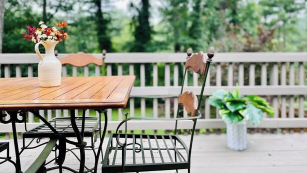 An outdoor wooden table with a white vase of flowers, a metal chair with a heart-shaped backrest, and a white fence in the background. - Bohemian Paradise | Near Downtown | Monthly Stays! (Asheville)