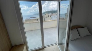 A bedroom with a sliding glass door leading to a balcony with a view of the sea and buildings. - Fenix Apartments (Bar)