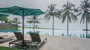 A poolside area with lounge chairs and a large umbrella, overlooking a beach with palm trees and the ocean.