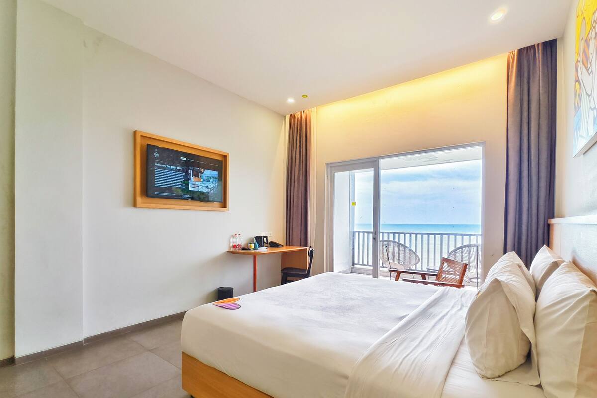 A hotel room with a large bed, a desk with a computer, and a view of the sea through a sliding door.