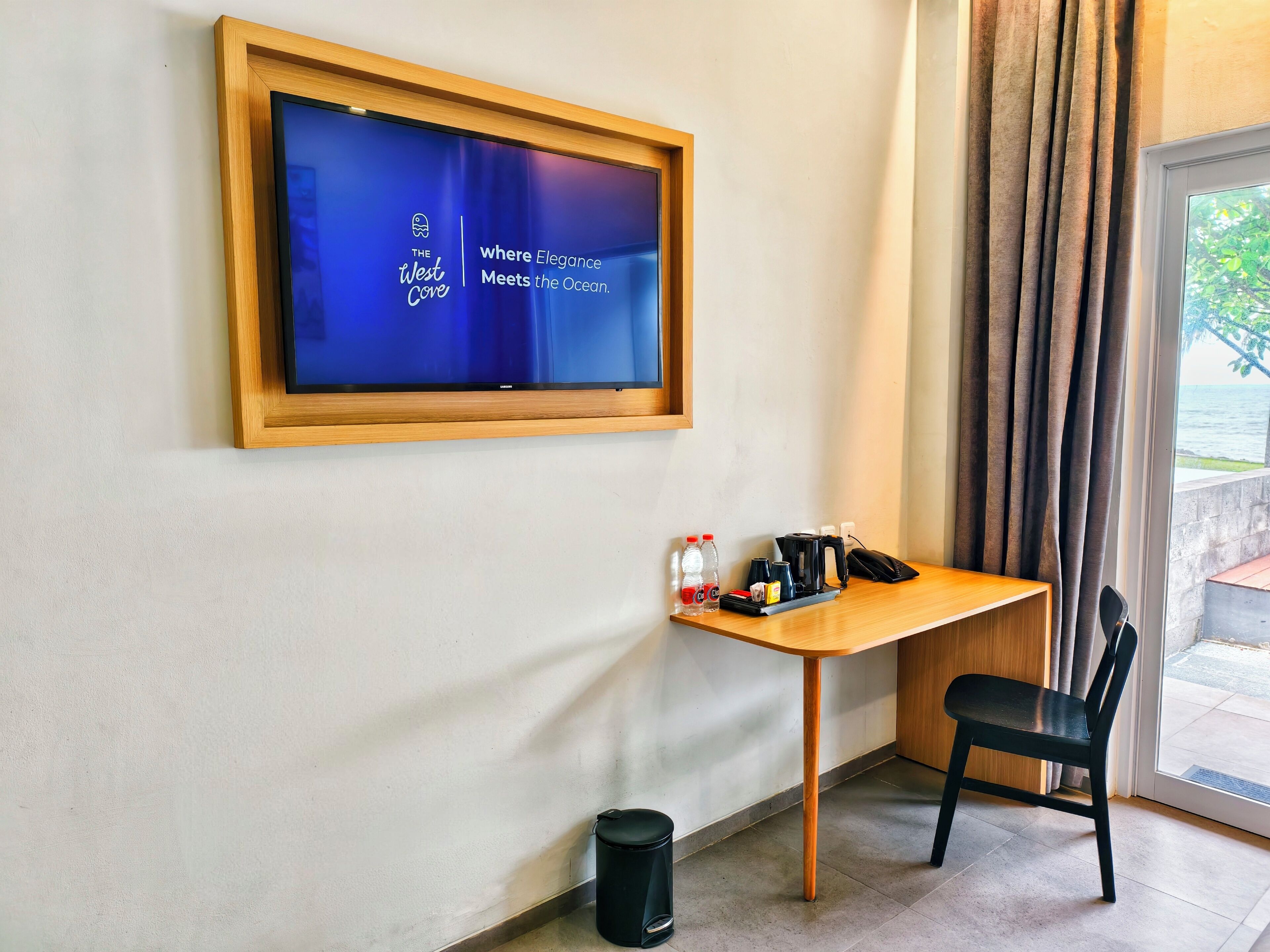 Une chambre d’hôtel avec un téléviseur affichant le logo de « The West Core », une petite table en bois avec une chaise, une poubelle et une vue sur l’océan par une fenêtre munie de rideaux.