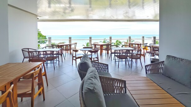 A spacious outdoor seating area with a view of the ocean, featuring wooden tables and chairs, and a cushioned seating area.