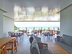 A spacious outdoor seating area with a view of the ocean, featuring wooden tables and chairs, and a cushioned seating area.
