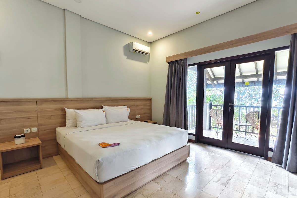 A hotel room with a large bed, a nightstand, an air conditioning unit, and a sliding glass door leading to an outdoor area with chairs.