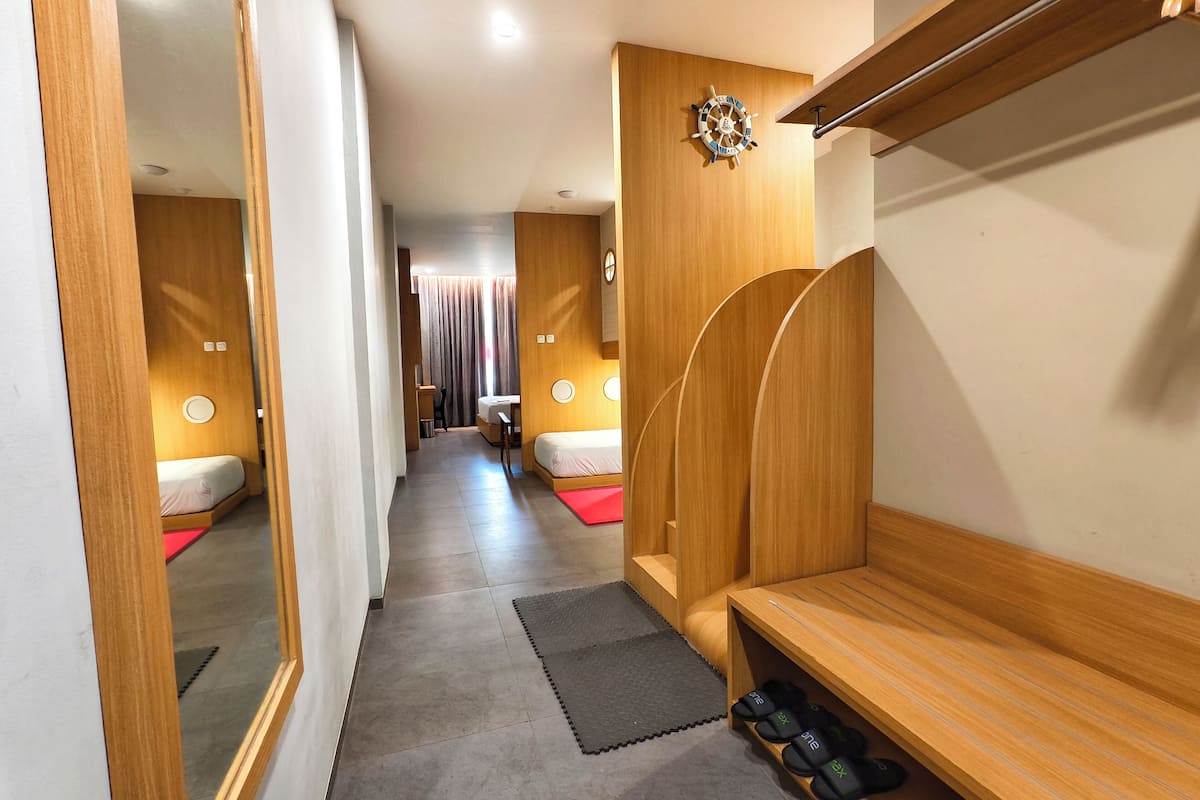A hotel room with a wooden vanity, a mirror, a bed with a red mat, a bench, and a shelf with slippers.
