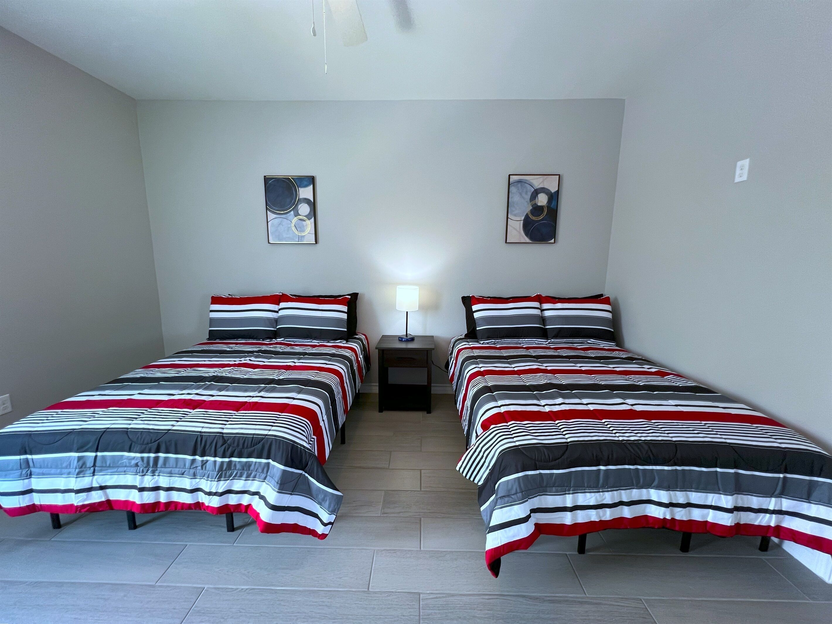 Ein Zimmer mit zwei Betten, jeweils mit gestreifter Bettwäsche in Rot, Weiß und Grau. Zwei Bilder hängen an der Wand, ein kleiner Tisch mit einer Lampe und ein Deckenventilator.