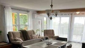 Ein geräumiges Wohnzimmer mit einem großen runden Tisch, bequemen Sitzgelegenheiten und großen Fenstern mit Blick ins Freie.