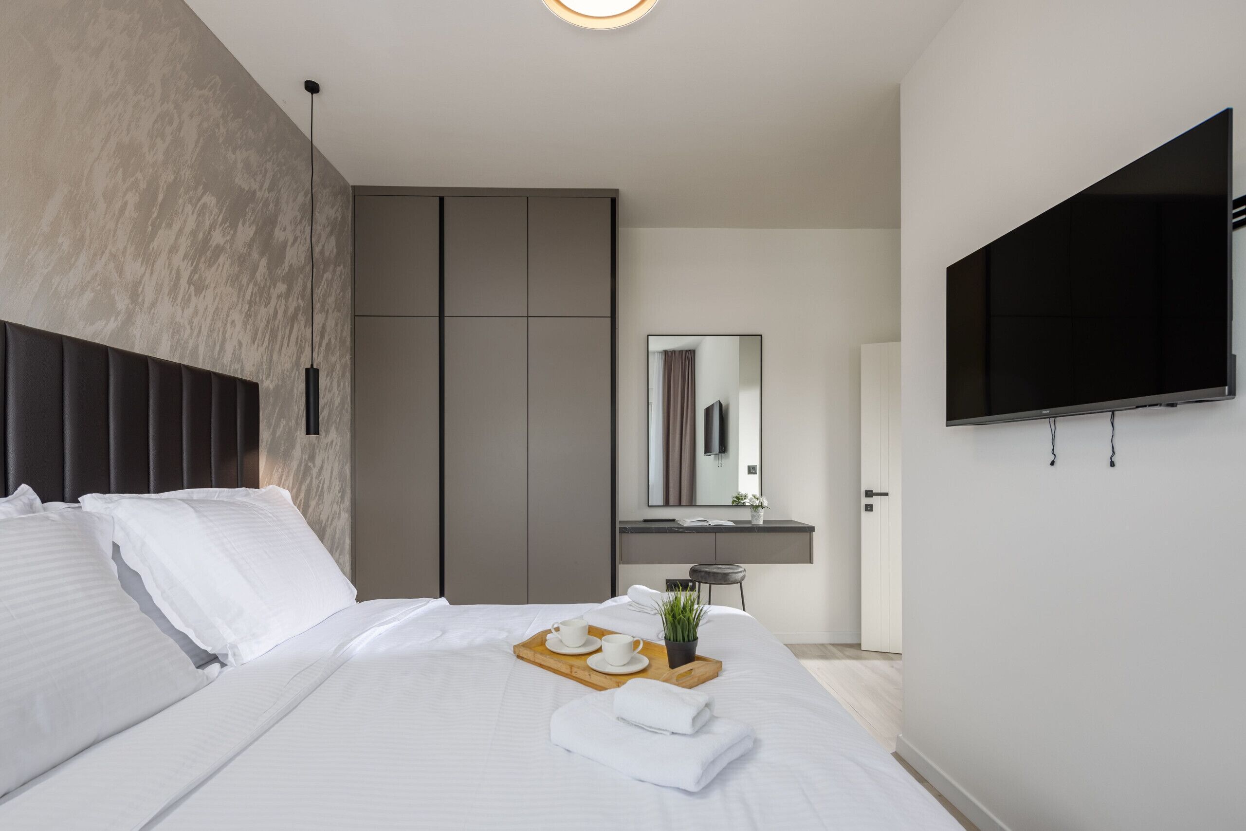 A modern hotel room with a large bed, a bedside table with a tray of breakfast items, a TV mounted on the wall, and a mirror above the sink.