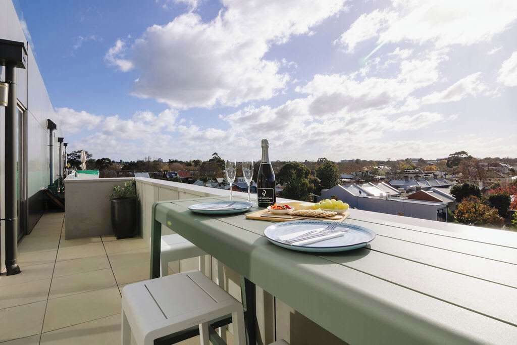 A balcony with a table set for a meal, a bottle, and a glass. The balcony overlooks a residential area with houses and trees.