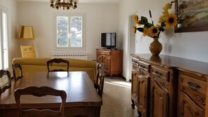 Une salle à manger avec une table et des chaises en bois, un téléviseur posé sur un meuble en bois, un radiateur et une fenêtre donnant sur des arbres.