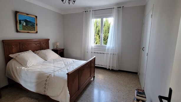 Une chambre à coucher avec un lit en bois, une table de chevet, une lampe, une fenêtre avec des rideaux et un radiateur.