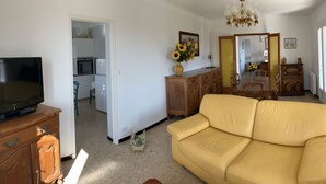 Un salon avec un canapé, un téléviseur posé sur un meuble en bois, une table à manger avec des chaises, et une vue sur le balcon extérieur.