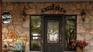 A stone house with a dark front door, a welcome sign, a rocking chair, and a flower pot. - Luxury in Wimberley | Pool+Pickleball+Party Barn (Wimberley)