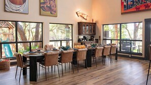 A spacious dining area with a large table and chairs, large windows, and a view of the outdoors. - Luxury in Wimberley | Pool+Pickleball+Party Barn (Wimberley)