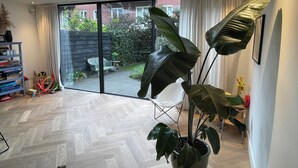 A spacious room with a large sliding glass door leading to a garden, a wooden herringbone floor, and a potted plant in the center. - Heerlijk Ruim Familie Appartement Vlakbij de Jordaan (Amsterdam)