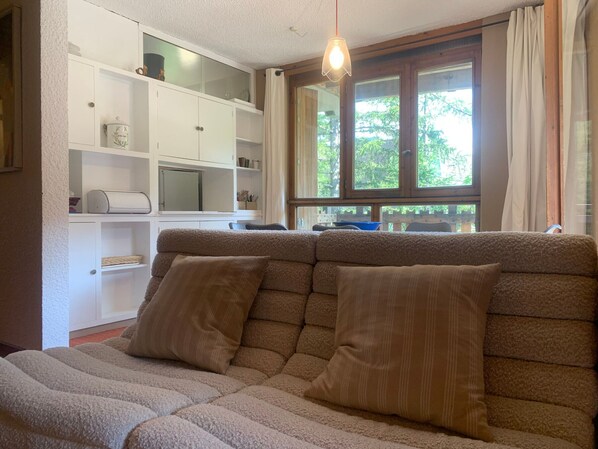A cozy living room with a sofa, a white cabinet with shelves, a television, and a window with curtains.