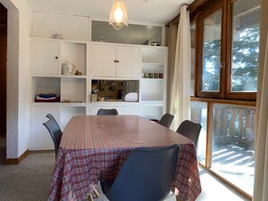 Ein Essbereich mit einem Tisch für sechs Personen, umgeben von Stühlen, einem weißen Schrank mit Einlegebögen für Geschirr und einem Fenster mit Blick auf eine Terrasse.