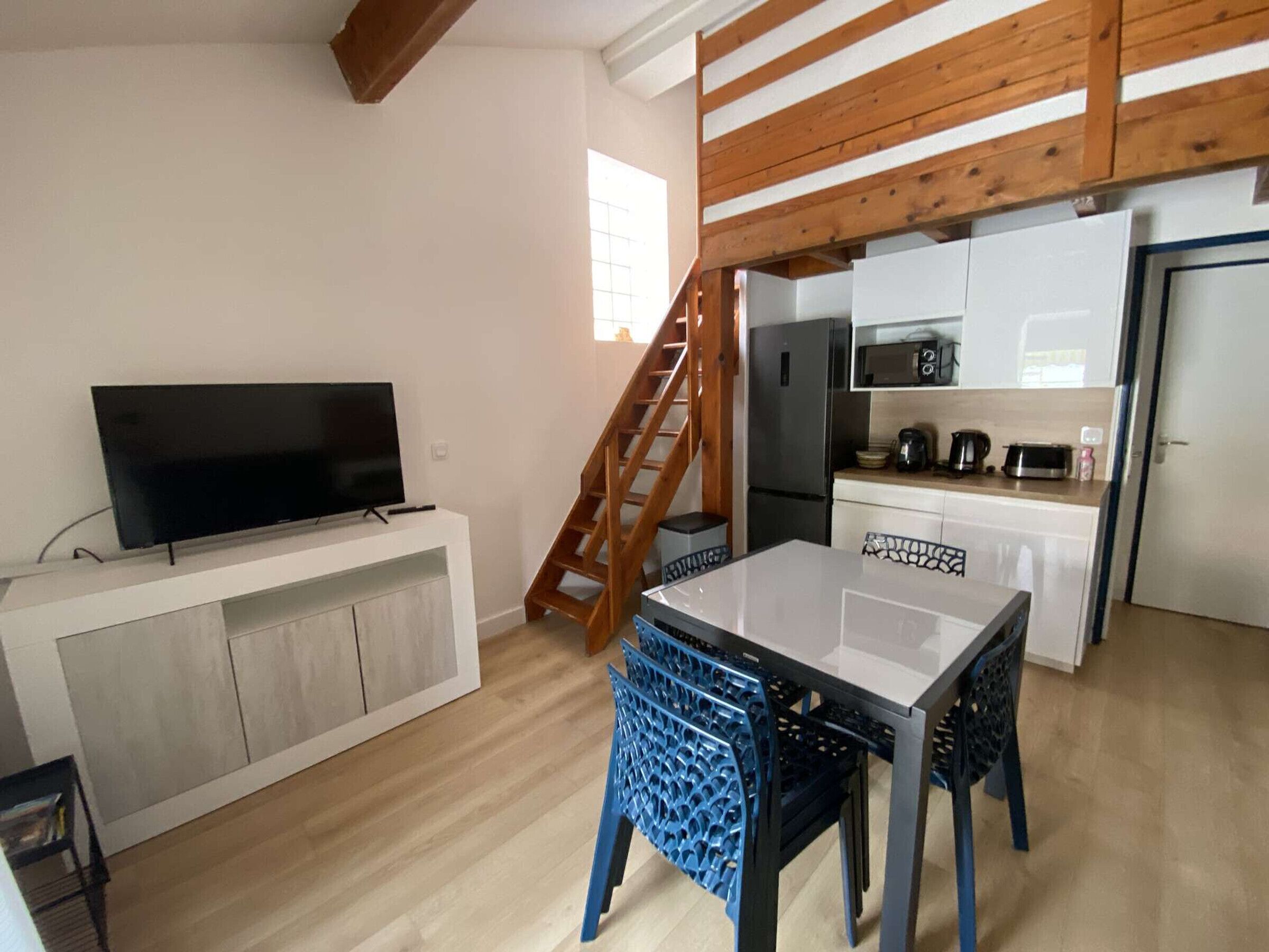 Un espace de vie compact comprenant un téléviseur posé sur un meuble blanc, un coin cuisine avec un four à micro-ondes et une table à manger avec des chaises bleues.