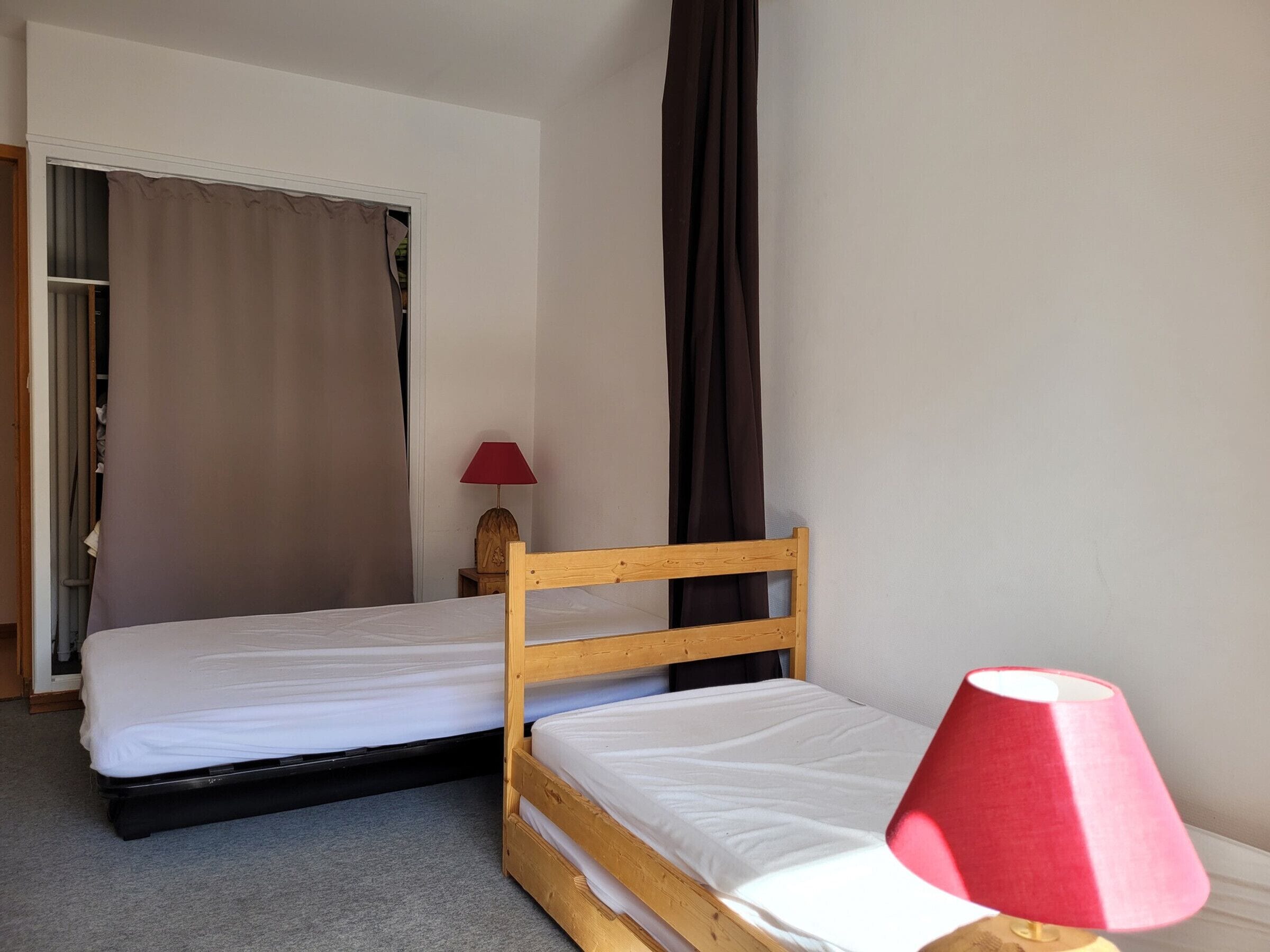 Un dormitorio pequeño y ordenado con dos camas individuales, cabecero de madera, una lámpara roja y un armario con cortina.