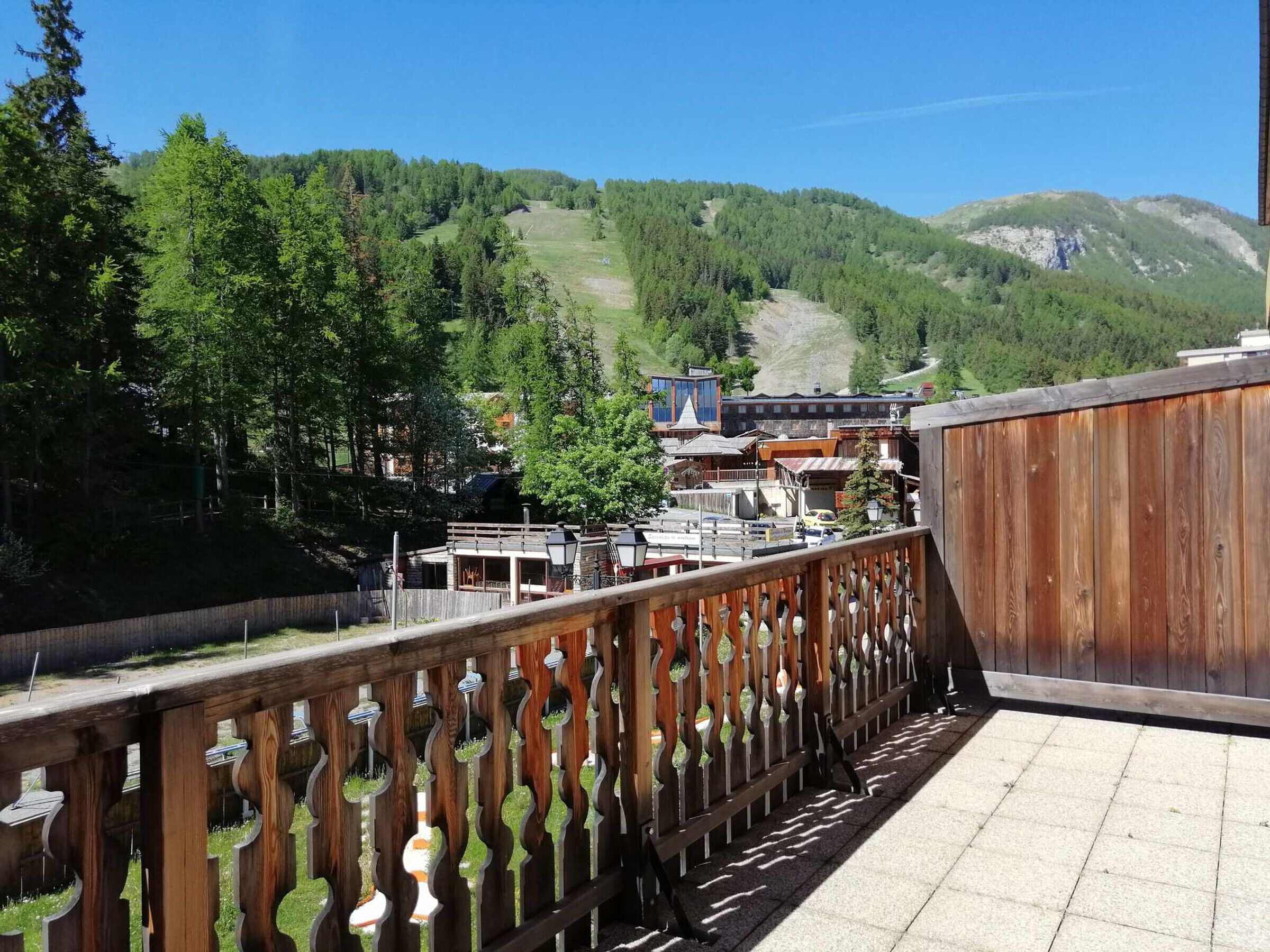 Een houten balkon met uitzicht op een bergachtig landschap, een gebouw met traditioneel ontwerp en een helderblauwe lucht.