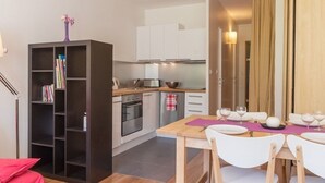 Una cocina compacta con electrodomésticos modernos, una zona de comedor con sillas blancas y una mesa dispuesta para dos personas, y una estantería llena de libros.