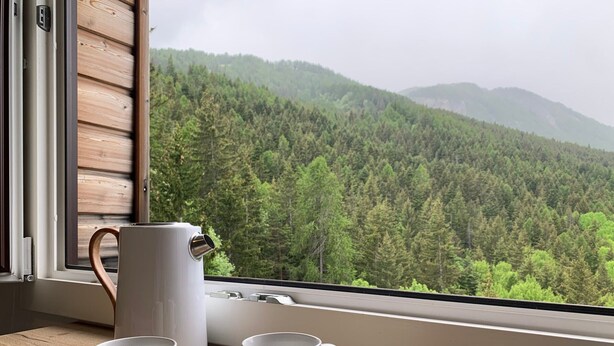 A wooden table with two white mugs and a white pitcher by a window overlooking a forested mountain.