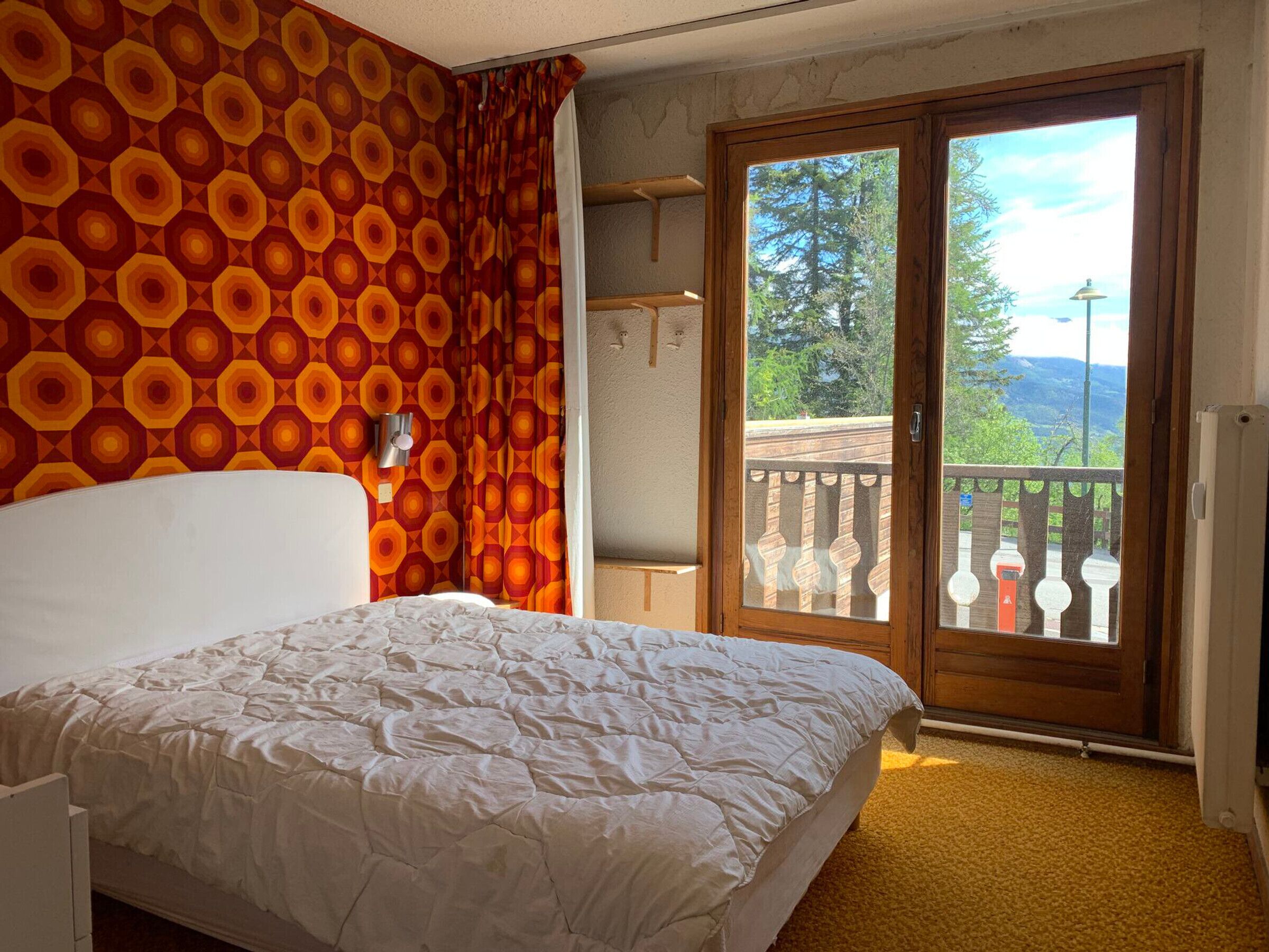 Ein Schlafzimmer mit Bett, Wand mit Muster, einer Schiebetür zu einem Balkon und Blick auf Bäume und Berge.